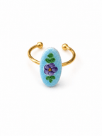 A gold-plated open cuff ring featuring a light blue oval stone hand-painted with a purple flower and green leaves, set against a plain white background.