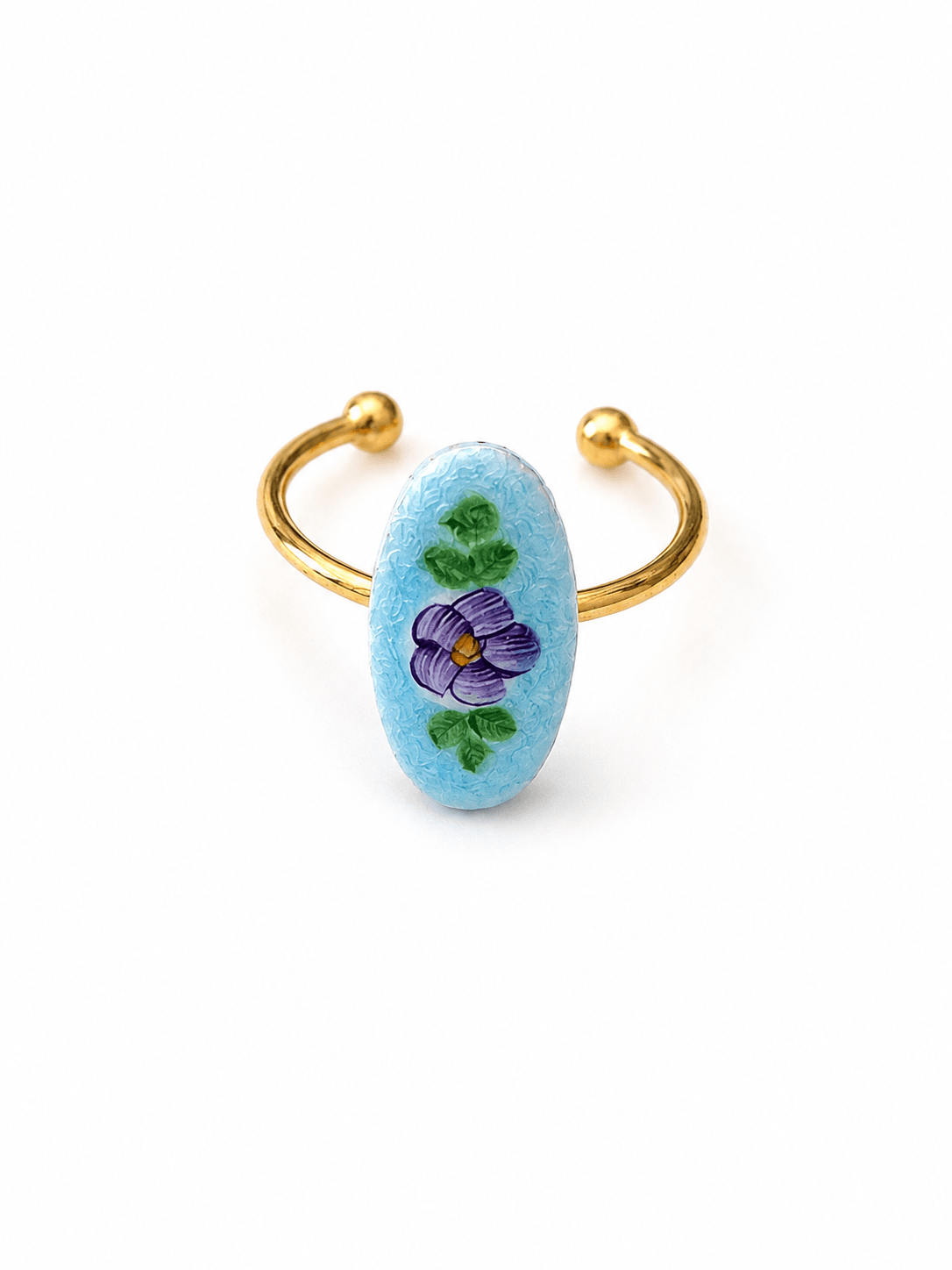 A gold-plated open cuff ring featuring a light blue oval stone hand-painted with a purple flower and green leaves, set against a plain white background.