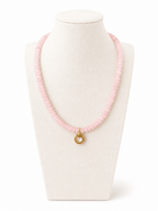 A pink beaded necklace with a small gold pendant featuring a clear round stone, displayed on a cream-colored jewelry bust against a white background.