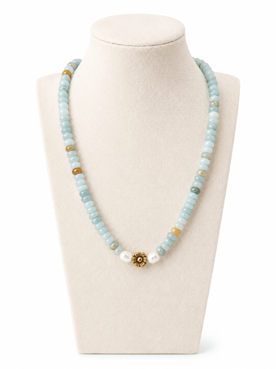 A beaded necklace displayed on a cream jewelry bust. The necklace features light blue stone beads, small white beads, a few tan/gold beads, and a central gold-toned floral or sunburst charm with clear stones.