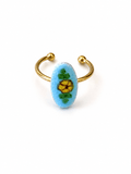 A gold-plated open cuff ring featuring a light blue oval bezel set with a yellow flower and green leaves, shown on a plain white background.