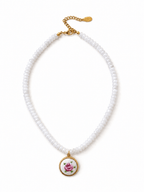 A white beaded necklace with a gold-tone chain extender and a round pendant featuring a pink rose on a white background with green leaves.