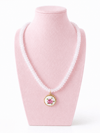 A white beaded necklace with a round pendant featuring a pink cartoon character on a white background with a gold border, displayed on a light pink jewelry bust against a white background.