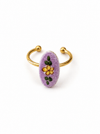 A gold-plated open cuff ring featuring a textured lavender purple oval centerpiece decorated with a yellow flower and green leaves, set against a plain white background.