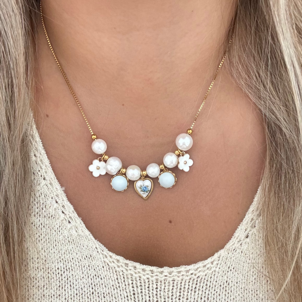 Model wearing the Freya Necklace, an 18K PVD gold-plated chain with baroque pearls, a white flower, a pastel blue glass pearl, and a blue rose heart ceramic charm. 17 inches.
