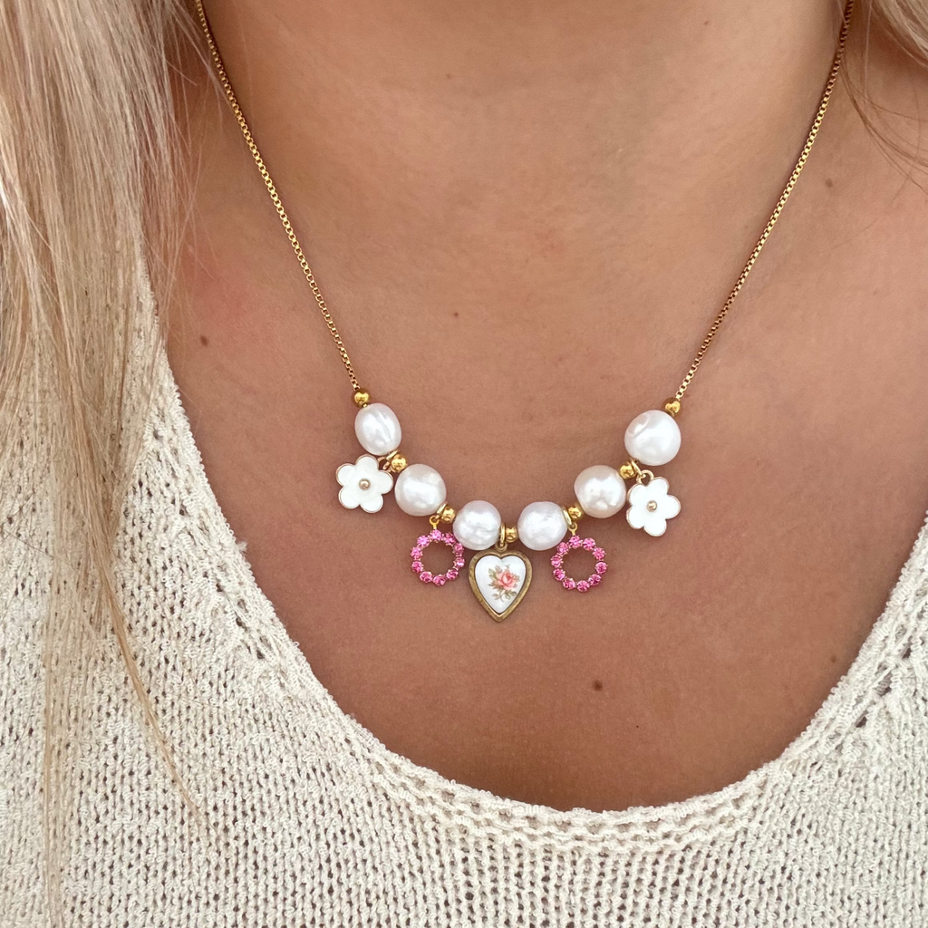Model wearing The Aria Necklace featuring an 18K PVD gold plated box chain with baroque natural pearls, dainty white flowers, a vintage Austrian rhinestone, and a pink floral heart ceramic set on a brass pendant. 17 inches in length.