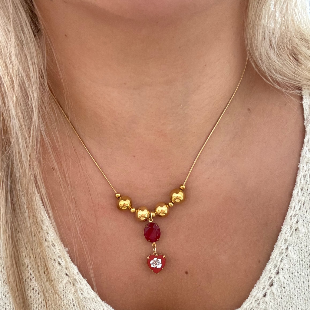 Model wearing The Inez Necklace featuring an 18K PVD gold plated snake chain with spacer beads, a red oval stone, and a vintage heart drop set on brass. 17 inches in length.
