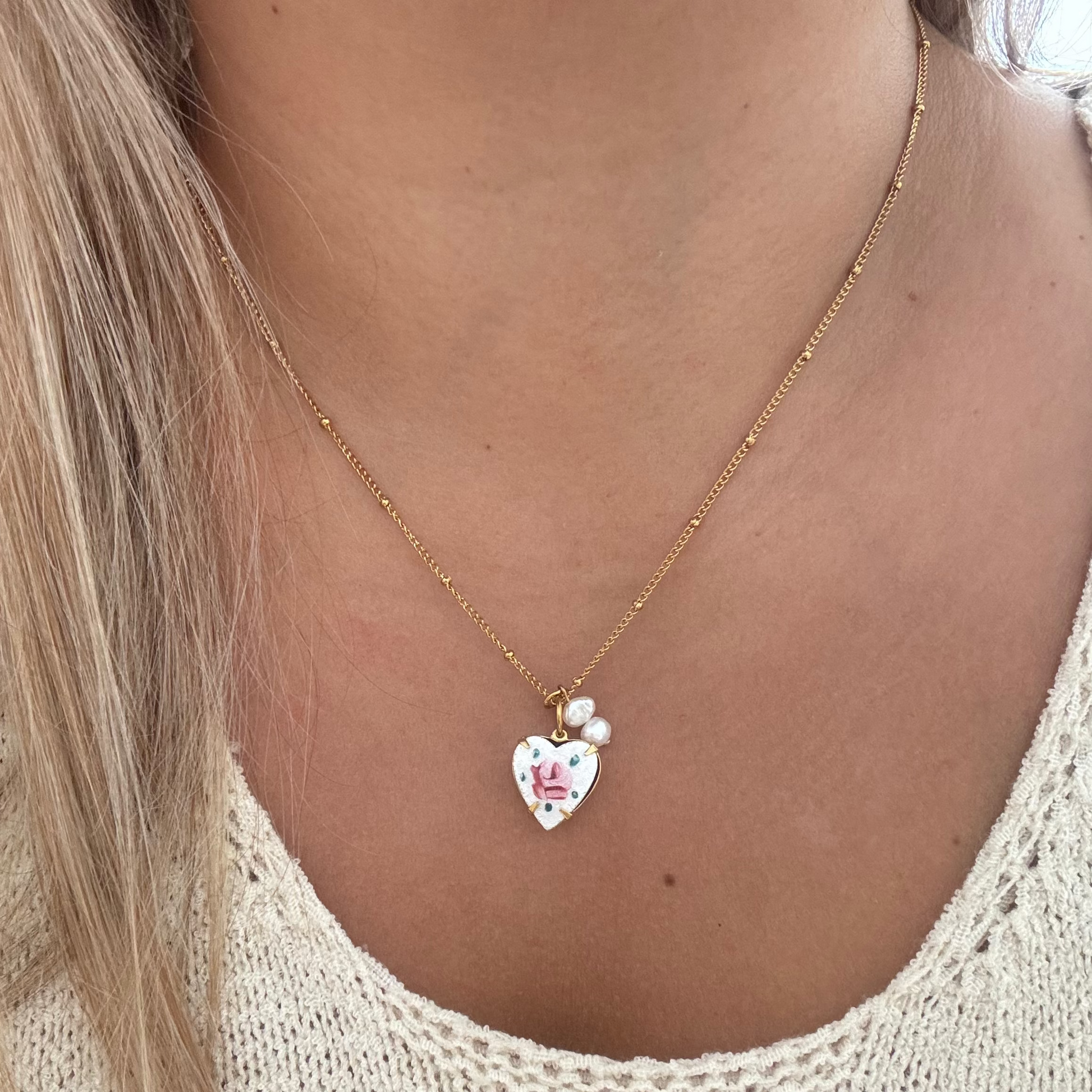Model wearing The Rosina Necklace featuring an 18K PVD gold-plated beaded Figaro chain with a spring ring lock, hand-painted rose on a heart guilloche, and two handmade baroque pearl drops. 17 inches in length