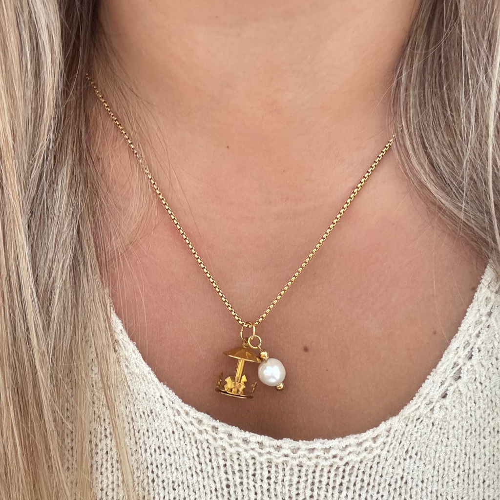 Model wearing The Gertrude Necklace featuring an 18K PVD gold plated cross chain with a brass spinning merry-go-round charm and a handmade freshwater pearl accent. A whimsical statement piece.