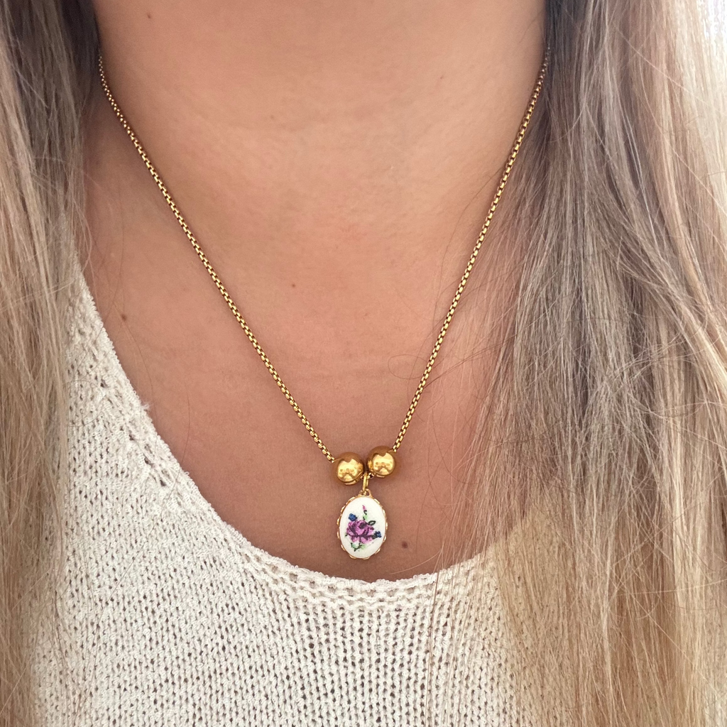 Model wearing The Hilda Necklace featuring an 18K PVD gold plated cross chain with a purple flower needlework design set on brass, accented by two gold-plated spacer beads. 17 inches in length