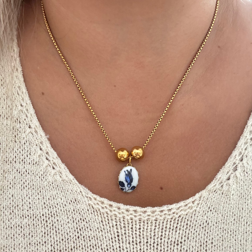 Model wearing The Phylis Necklace featuring an 18K PVD gold plated cross chain with a blue tulip design on oval ceramic, set in gold or brass, and accented by two gold-plated spacer beads. 17 inches in length.