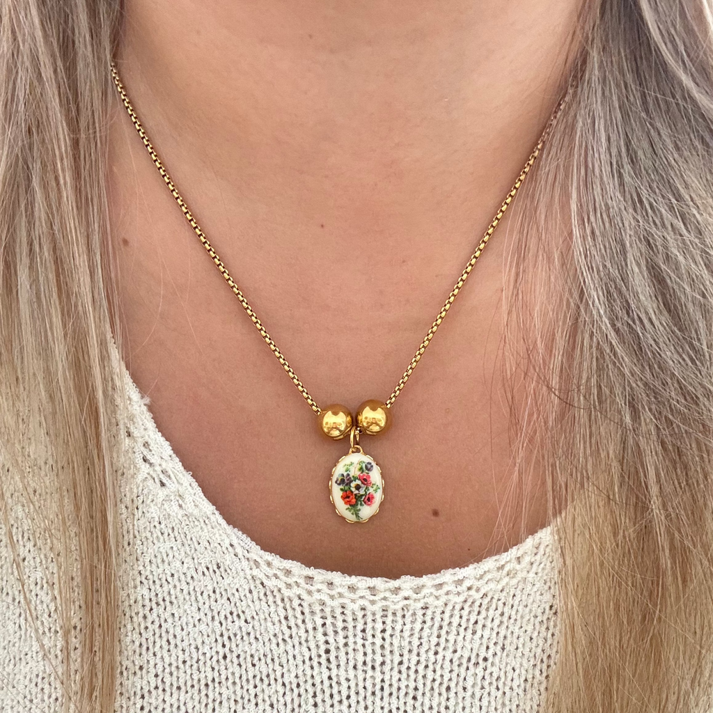 Model wearing The Lily Necklace featuring an 18K PVD gold plated cross chain with a poppy flower bouquet on ceramic, set in a gold-plated frame with two gold-plated spacer beads. 17 inches in length.