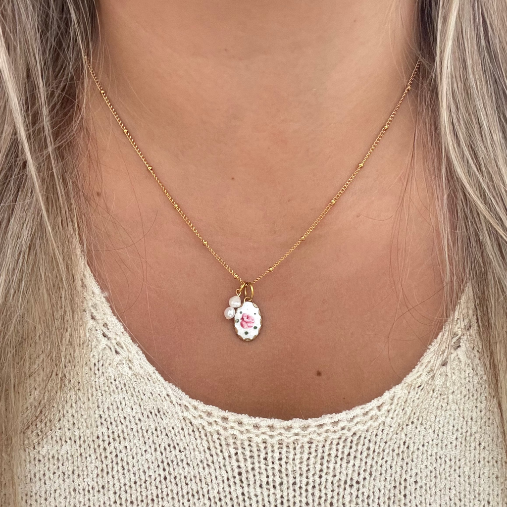Model wearing The Muriel Necklace featuring an 18K PVD gold-plated beaded Figaro chain with a spring ring lock, hand-painted rose on an oval guilloche set on brass, and two handmade baroque pearl drops. 17 inches in length.