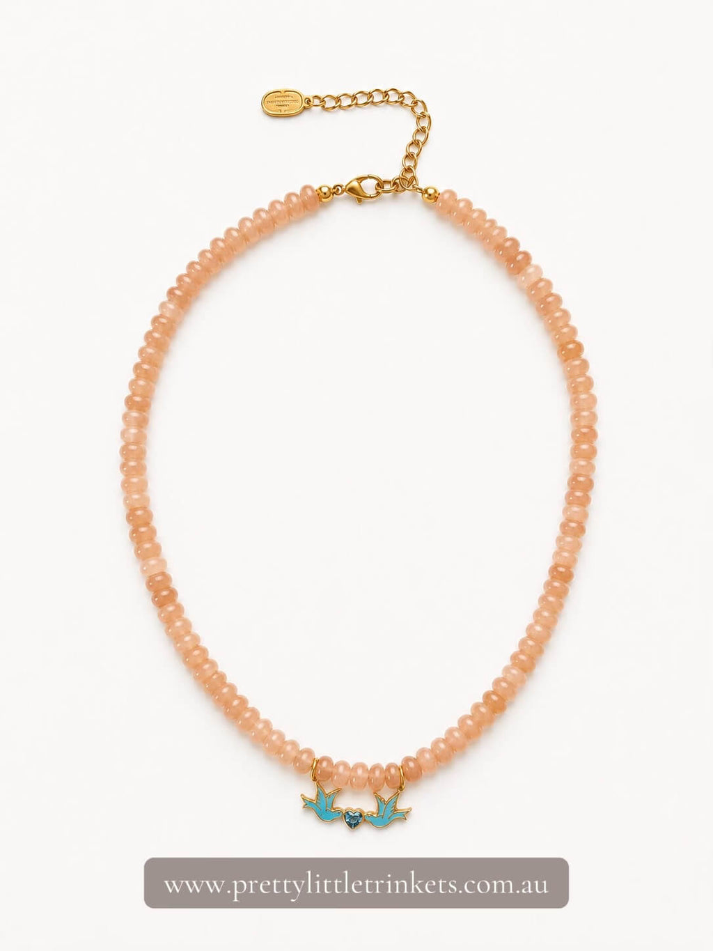 A peach-colored beaded necklace with a gold-tone chain extender and two small turquoise bird charms, displayed on a clean white background with a brand watermark at the bottom.