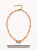 A peach-colored beaded necklace with a gold-tone chain extender and two small turquoise bird charms, displayed on a clean white background with a brand watermark at the bottom.