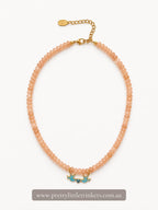 A peach-colored beaded necklace with a gold-tone chain extender and two small turquoise bird charms, displayed on a clean white background with a brand watermark at the bottom.