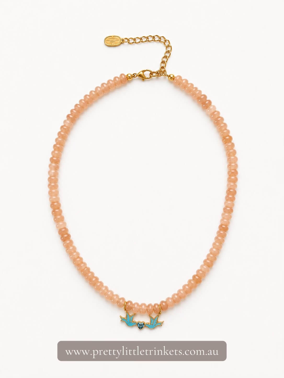 A peach-colored beaded necklace with a gold-tone chain extender and two small turquoise bird charms, displayed on a clean white background with a brand watermark at the bottom.