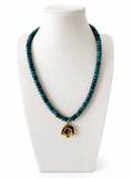 A beaded necklace with dark teal-blue faceted beads and a gold rose-shaped pendant, displayed on a white fabric jewelry bust against a white background.
