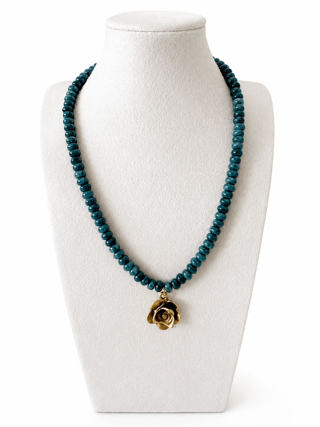 A beaded necklace with dark teal-blue faceted beads and a gold rose-shaped pendant, displayed on a white fabric jewelry bust against a white background.