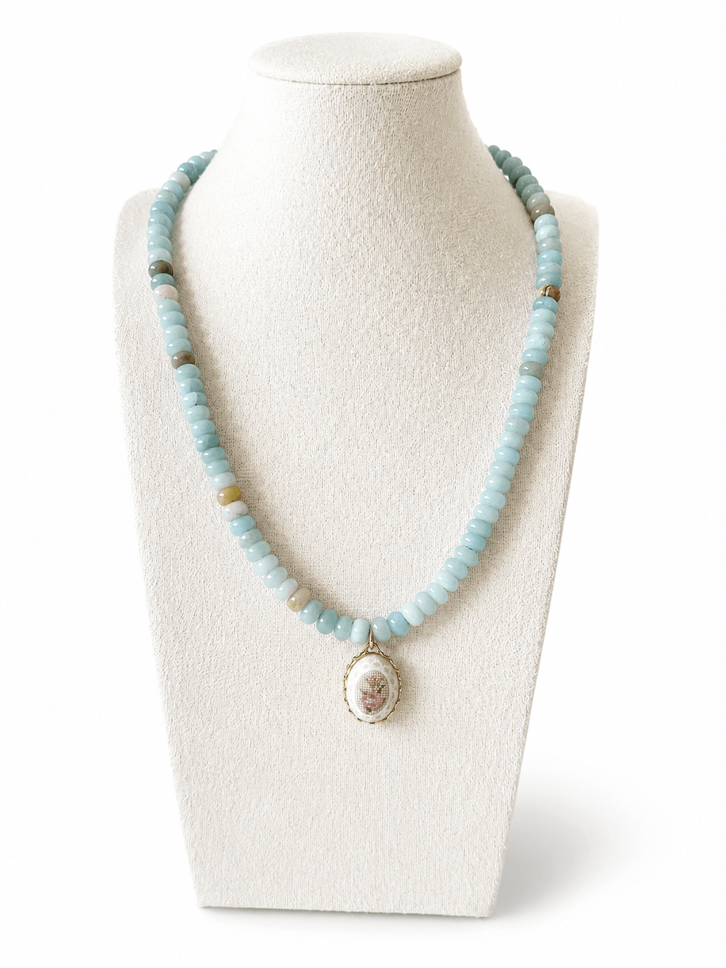 A light blue beaded necklace with a vintage-style oval pendant featuring a pink floral design and gold trim, displayed on a cream-colored mannequin bust against a white background.
