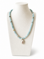 A light blue beaded necklace with a vintage-style oval pendant featuring a pink floral design and gold trim, displayed on a cream-colored mannequin bust against a white background.