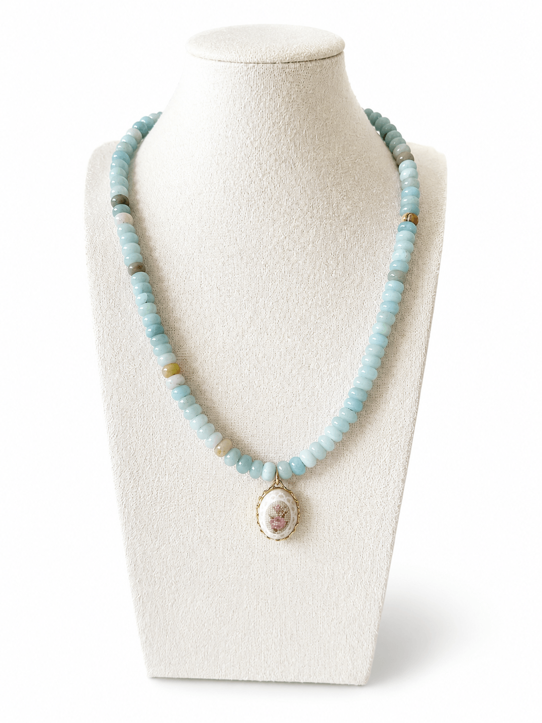 A light blue beaded necklace with a vintage-style oval pendant featuring a pink floral design and gold trim, displayed on a cream-colored mannequin bust against a white background.
