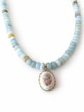 A beaded necklace featuring light blue and white round beads with a central oval pendant showcasing a pink and white cross-stitch style design framed in gold.