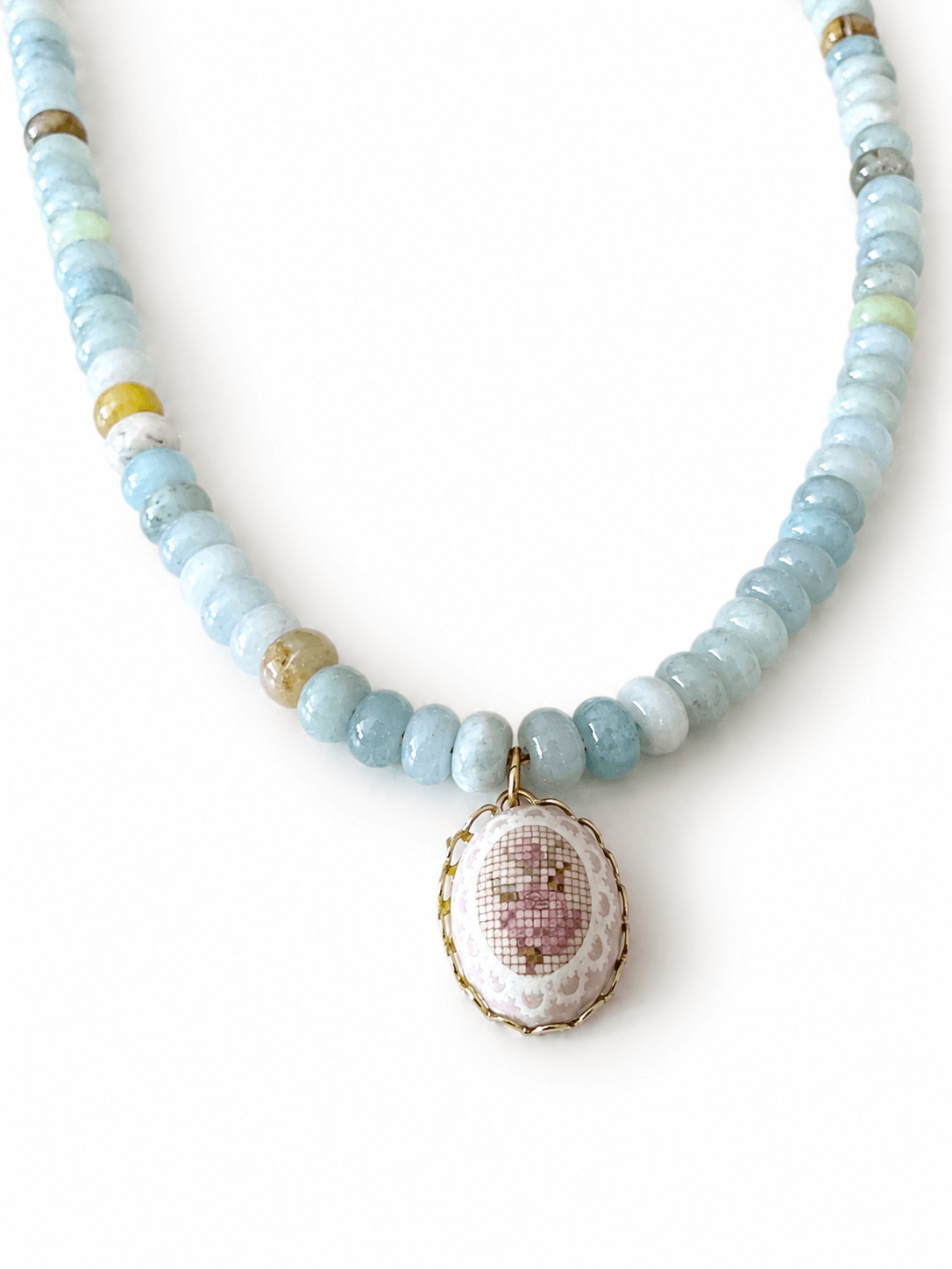 A beaded necklace featuring light blue and white round beads with a central oval pendant showcasing a pink and white cross-stitch style design framed in gold.