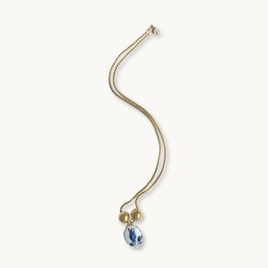 Phylis Necklace - 18K gold-plated cross chain with an oval ceramic pendant featuring a blue tulip design, set in gold or brass, with gold-plated spacer beads, 17 inches in length.