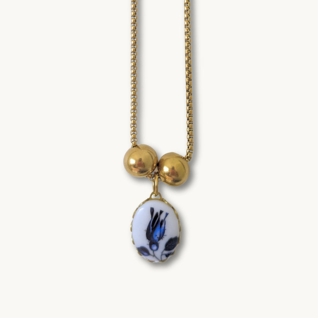 Close-up of the Phylis Necklace showcasing its delicate blue tulip ceramic pendant on a gold-plated cross chain.