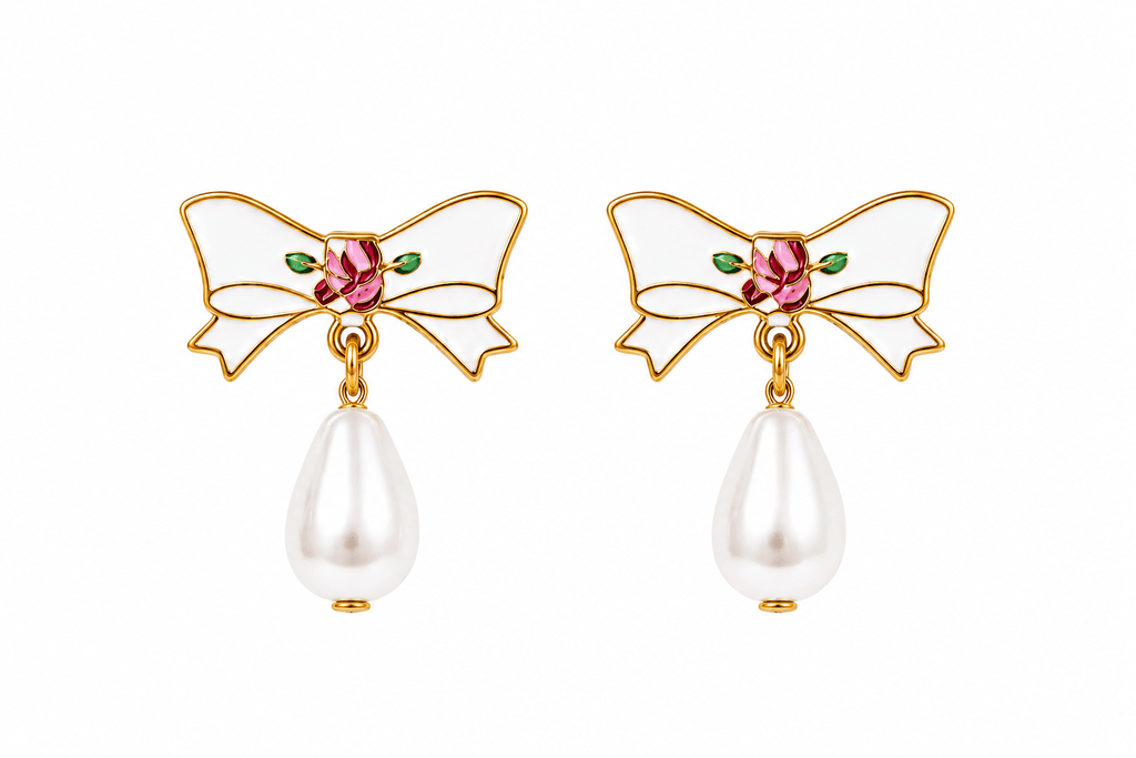 A pair of gold bow earrings with white enamel, a pink rhinestone center, small green enamel leaves, and a dangling white pearl teardrop below each bow.