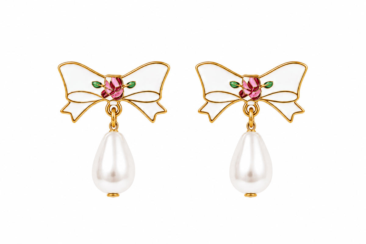 A pair of gold bow earrings with white enamel, a pink rhinestone center, small green enamel leaves, and a dangling white pearl teardrop below each bow.