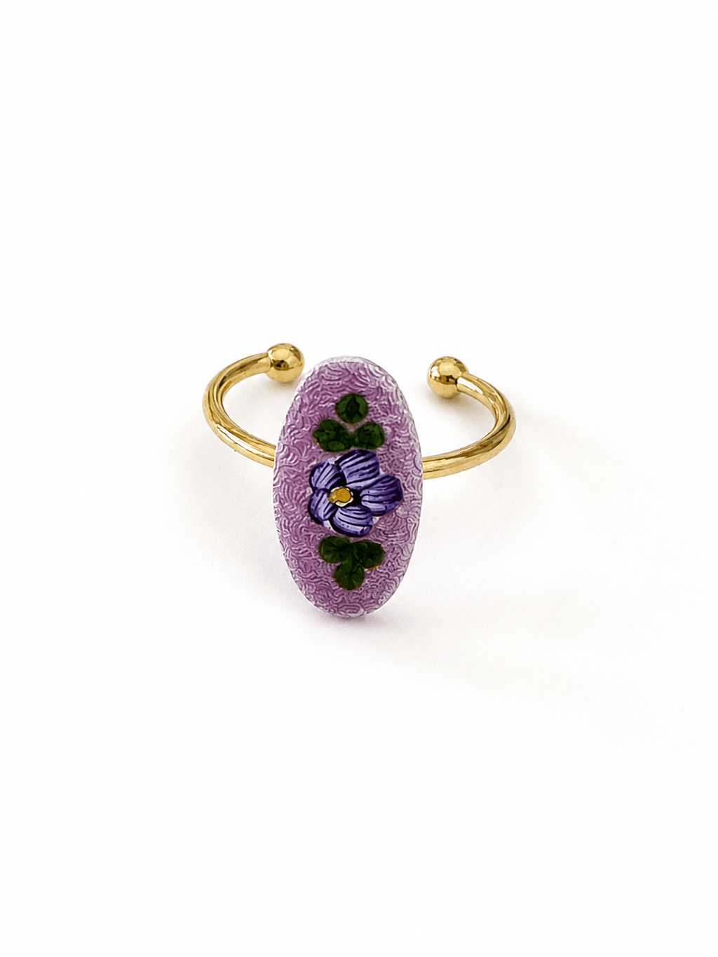 A gold-plated open cuff ring featuring a textured, oval-shaped lavender purple stone inlaid with a hand-painted purple pansy and green leaves, set against a plain white background.