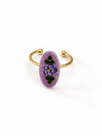 A gold-plated open cuff ring featuring a textured, oval-shaped lavender purple stone inlaid with a hand-painted purple pansy and green leaves, set against a plain white background.