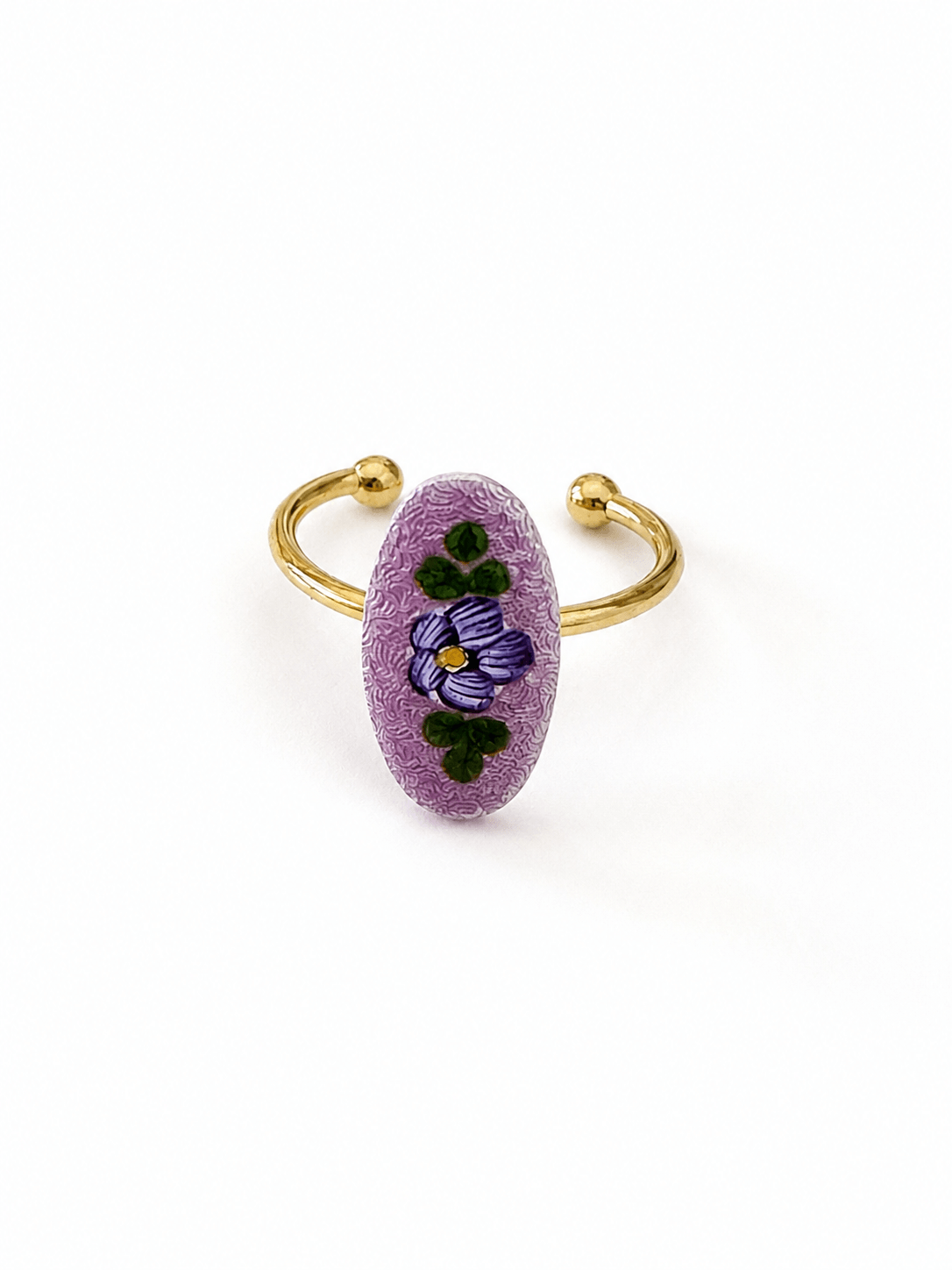 A gold-plated open cuff ring featuring a textured, oval-shaped lavender purple stone inlaid with a hand-painted purple pansy and green leaves, set against a plain white background.