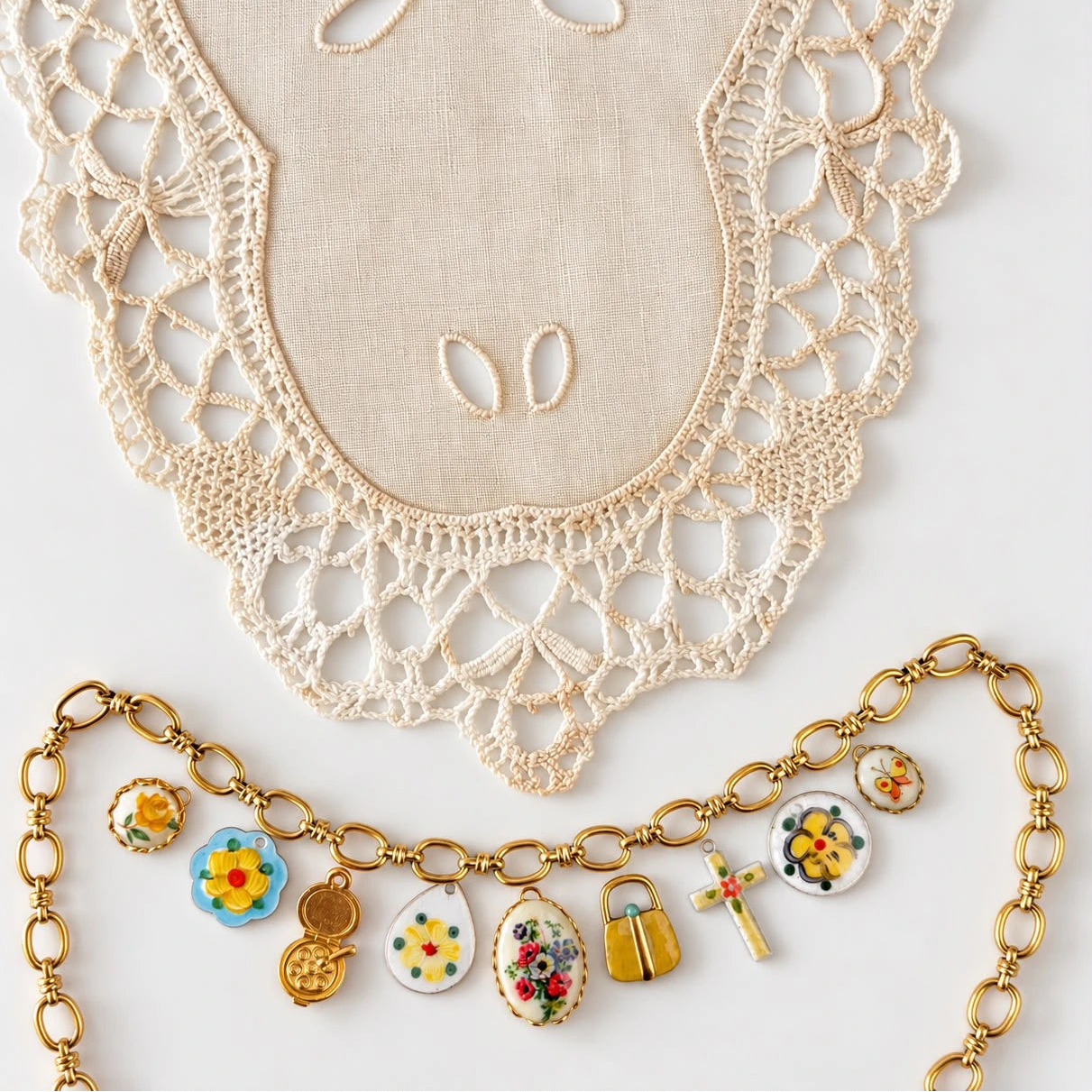 A gold-tone chain necklace with colorful enamel charms, displayed on a light beige fabric backdrop with a matching lace textile above it.